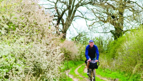 A man cycles towards us along an overgrown country lane, flowers blooming on the hedges surrounding him