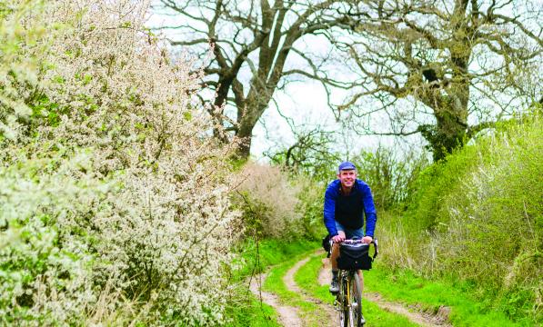 A man cycles towards us along an overgrown country lane, flowers blooming on the hedges surrounding him