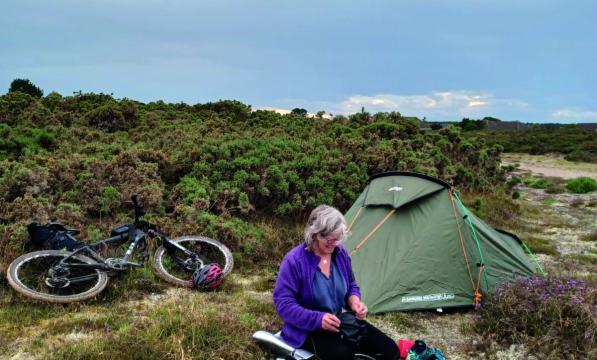 A woman sits amongst heather next to a pitched tent, her bicycle laying behind her