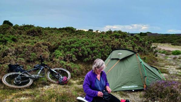 A woman sits amongst heather next to a pitched tent, her bicycle laying behind her