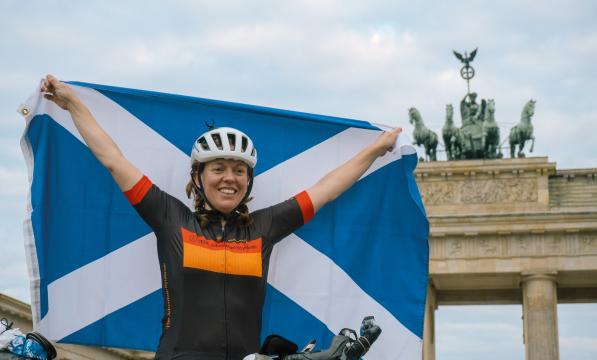 A woman stands holding a Scottish flag behind her while posing in cycling gear in front of the Brandenburg gates in Berlin, Germany