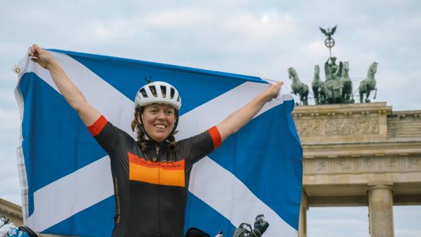 A woman stands holding a Scottish flag behind her while posing in cycling gear in front of the Brandenburg gates in Berlin, Germany