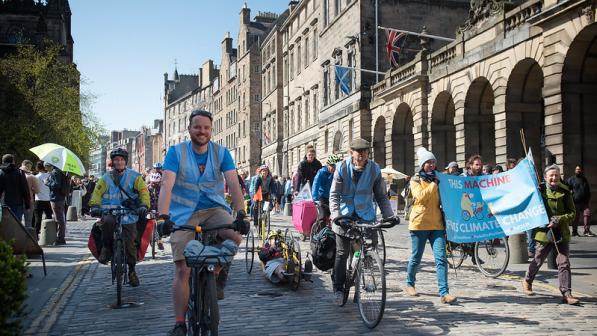Many people are cycling down a cobbled street with grand buildings, including people on standard two-wheeled cycles and a three-wheeled handcycle. Two people are carrying a banner with a bicycle on it that reads 'This machine fights climate change'.