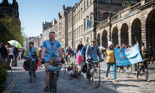 Many people are cycling down a cobbled street with grand buildings, including people on standard two-wheeled cycles and a three-wheeled handcycle. Two people are carrying a banner with a bicycle on it that reads 'This machine fights climate change'.