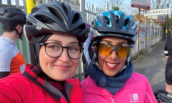 Two ladies pose for a selfie. They are wearing brightly coloured cycling jackets, glasses and helmets.