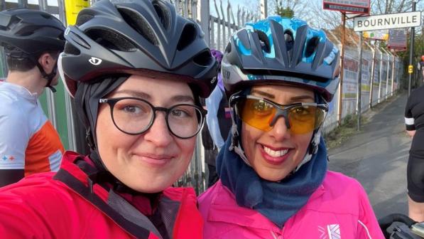 Two ladies pose for a selfie. They are wearing brightly coloured cycling jackets, glasses and helmets.