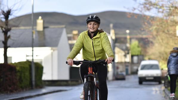 A girl wearing a yellow jacket and helmet is smiling as she cycles up the road. There are houses and hill in the background