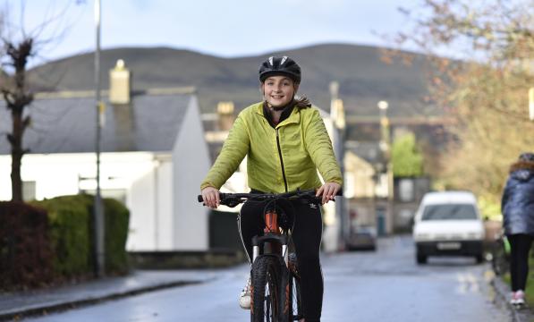 A girl wearing a yellow jacket and helmet is smiling as she cycles up the road. There are houses and hill in the background