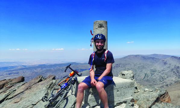 A young man sits at the summit of a mountain with his bicycle next to him. 