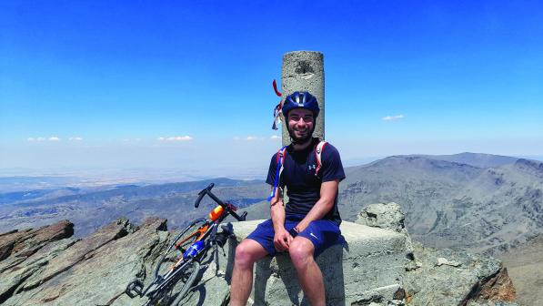A young man sits at the summit of a mountain with his bicycle next to him. 