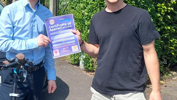 Two young men stand side by side in front of a garden hedge on a pavement. They are both holding a certificate