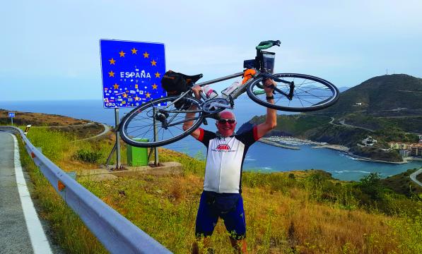 A older man wearing cycling clothes holds a road bicycle above his head in triumph along a coastal road. Behind him, a road sign with the EU flag on it surrounds the word 'Espana'.