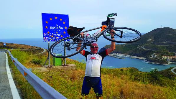 A older man wearing cycling clothes holds a road bicycle above his head in triumph along a coastal road. Behind him, a road sign with the EU flag on it surrounds the word 'Espana'.