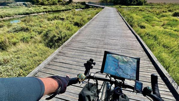 POV of a cyclist riding along a raised boardwalk above marshland. Their bike has a map attached to the handlebars and pannier bags on the forks