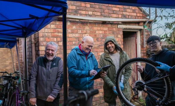 Four men are seen standing under a marquee. They are laughing amongst themselves and smiling at the camera, one holds a bicycle wheel and another holds a clipboard
