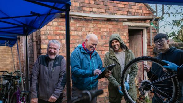 Four men are seen standing under a marquee. They are laughing amongst themselves and smiling at the camera, one holds a bicycle wheel and another holds a clipboard