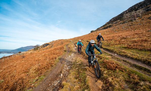 Three mountain bikers ride along a bridleway on a hill side. In the background there is a lake and hillside.