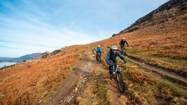 Three mountain bikers ride along a bridleway on a hill side. In the background there is a lake and hillside.