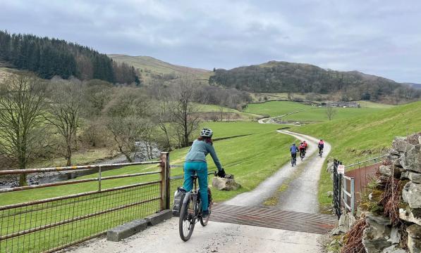 Five cyclists ride along a gravel road weaving its way through a valley, they are cycling over a cattle grid