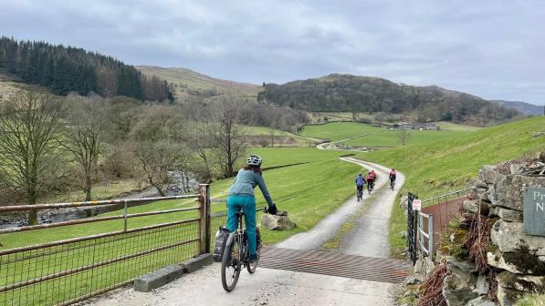 Five cyclists ride along a gravel road weaving its way through a valley, they are cycling over a cattle grid