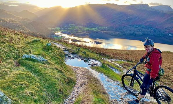 Man stands astride a mountain bike in front of a spectacular view of the sun setting behind a mountain and a trail heading down towards a lake