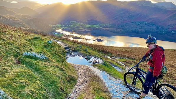 Man stands astride a mountain bike in front of a spectacular view of the sun setting behind a mountain and a trail heading down towards a lake