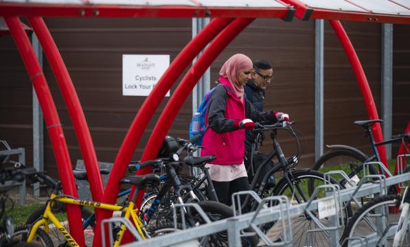 A woman pushes her bike into a bike rack in a red bike shelter. Man in background doing the same. 