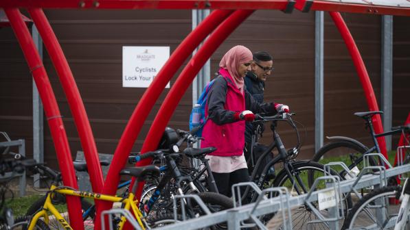A woman pushes her bike into a bike rack in a red bike shelter. Man in background doing the same. 