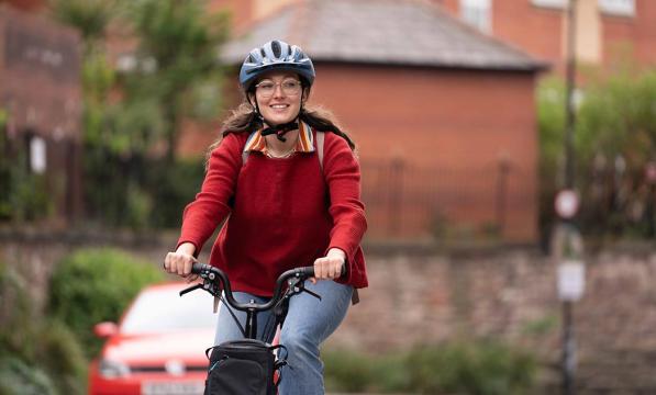 A woman cycles along a road. She is wearing a red knitted jumper, a pair of jeans, a helmet and a pair of glasses. She is smiling