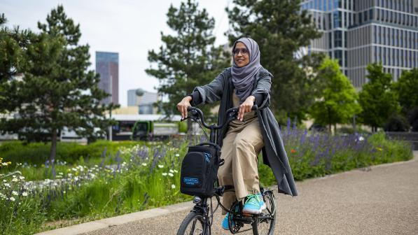 Woman pedals electric Brompton along cycle path with purple, white and yellow flowers and trees in background