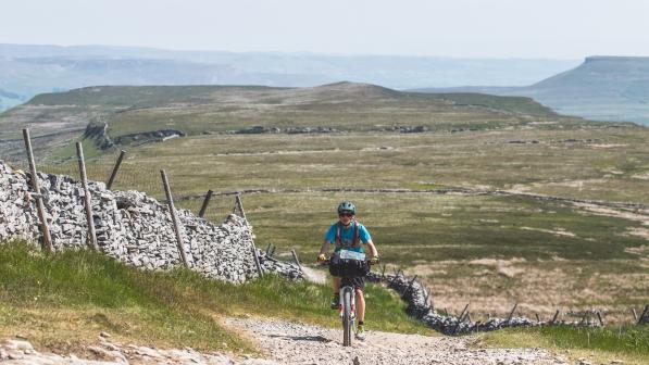 Woman cycles up a rough gravel track towards the camera, with an expansive moorland view behind her