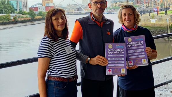 Three people standing side-by-side at Newcastle riverside. Two of them are holding certificates