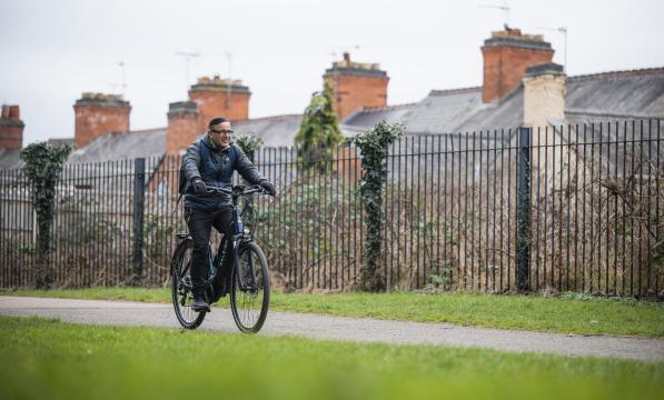 A man wearing glasses pedals a navy e-cycle through park in Leicester on a cloudy day
