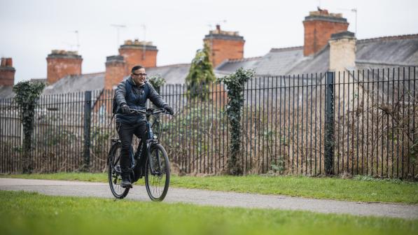 A man wearing glasses pedals a navy e-cycle through park in Leicester on a cloudy day