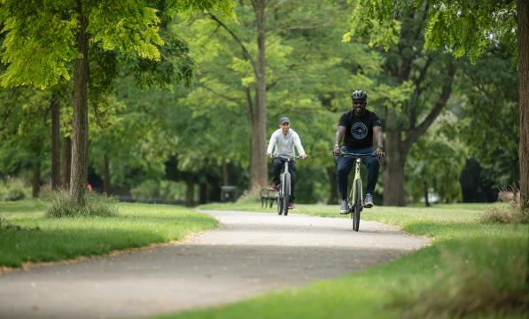 Two men pedal e-bikes through a park on a sunny day