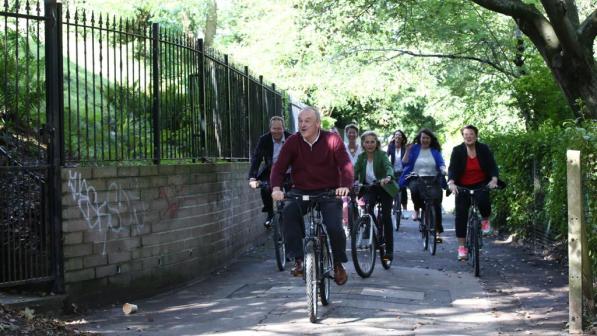 A group of people in normal clothes led by a man cycle down a path. There is a fence on the left with some greenery behind it