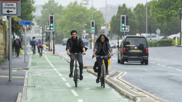 Two people ride side by side on a green cycle lane on a grey day in the city. Traffic lights on green in the background.