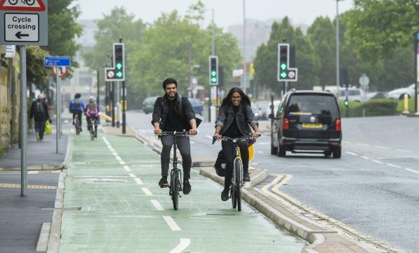 Two people ride side by side on a green cycle lane on a grey day in the city. Traffic lights on green in the background.
