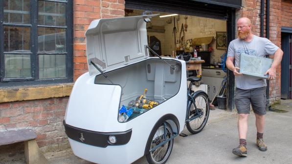 A stonemason wearing a grey t-shirt loads tools into a white e-cargo bike outside his workshop