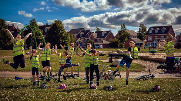 A group of children in hi-viz vests are jumping up in the air in a park next to their bikes on the ground