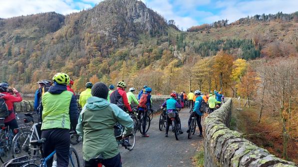 A group of people is cycling along a quiet country road with a stone wall running alongside. It's autumn and the leaves are all copper and red.