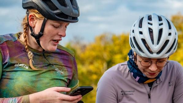 Two women wearing cycling jerseys and helmets. One is looking down, the other has a phone in her hand