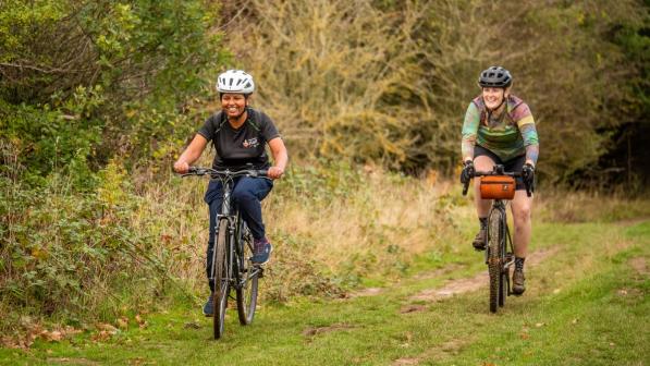 Two women are riding along a grassy path in the countryside. One is riding a hybrid bike, the other a gravel bike. They are quite muddy and are laughing