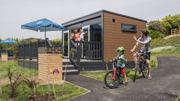 A family waving at each other, next to a glamping pod and on bikes. 