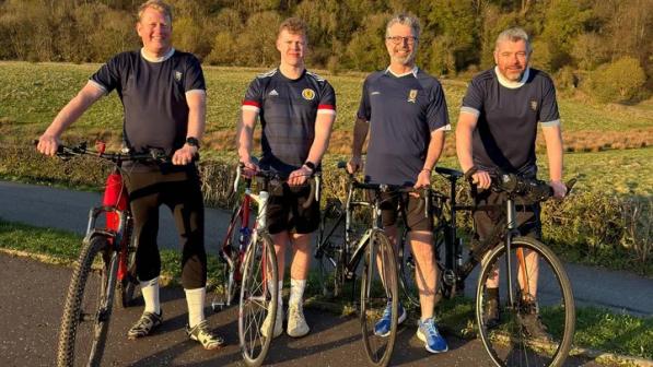 Four men are standing with their bikes on a paved path. They’re wearing Scotland football shirts and black shorts, one is wearing black tracksuit bottoms. Two bikes are hybrids, two are road bikes.