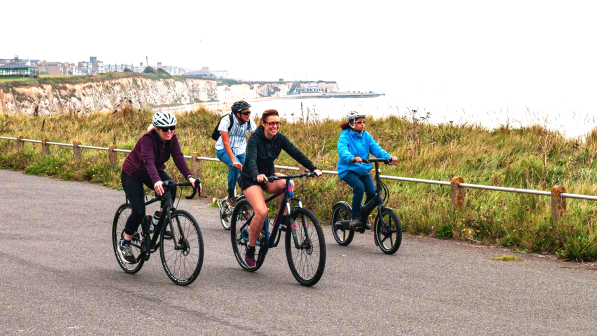Four people are cycling along a paved path on a clifftop with a seaside town in the background. They're on a mix of bikes, including road, hybrid and folding. They're wearing normal clothes.
