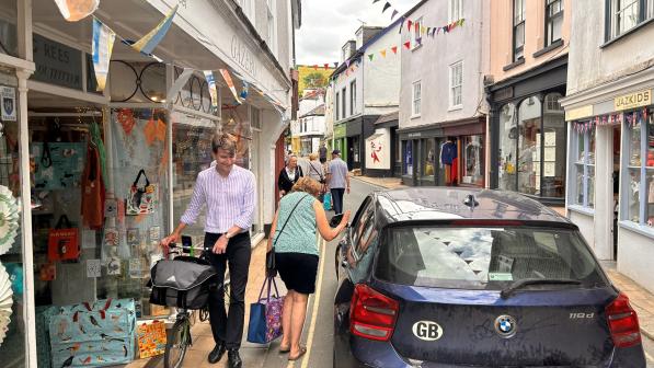 A narrow street in a quaint town, lined with small shops and decorated with colorful bunting. A man in a striped shirt is seen walking a folded bicycle on the sidewalk next to a shop with a window display of bags and accessories. A woman beside him is navigating around a parked car that is partially blocking the pathway. Other pedestrians can be seen walking further down the street. The atmosphere feels lively yet constrained due to the tight space shared by pedestrians and parked vehicles.