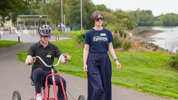 A man is cycling along a paved path on an adapted four-wheel bike, a woman in blue wide-legged trousers and a Cycling UK T-shirt is walking alongside him