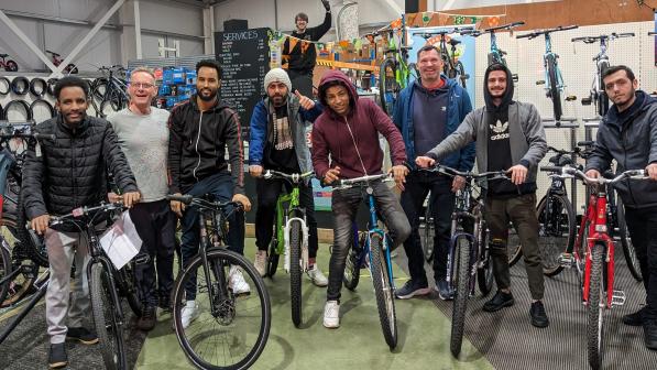 Group of nine individuals standing inside a bicycle shop, all smiling and holding bicycles. They are posing in front of a variety of bikes displayed on racks, with one person in the background standing on a platform, giving a thumbs-up. The group consists of a mix of ages, dressed in casual clothing, and they appear excited and ready to ride.