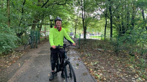 Matthew, wearing a bright green jacket and helmet, stands with his e-bike on a peaceful, tree-lined path, surrounded by lush greenery.